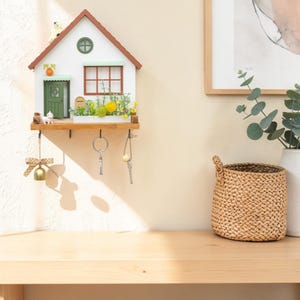 Puede incluir: Un llavero de pared de madera con forma de casa, fachada blanca, tejado marr&oacute;n y puerta verde. Incluye una ventana peque&ntilde;a, un buz&oacute;n y una jardinera con flores amarillas. Debajo cuelgan cuatro ganchos met&aacute;licos para las llaves.