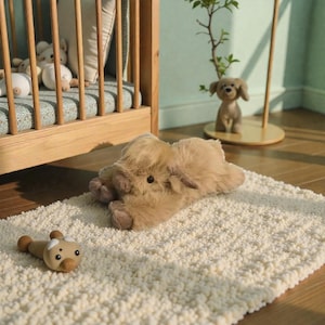 May include: A fluffy, light brown plush highland cow toy lying on a textured cream-coloured rug. Nearby, a small, smooth, tan-coloured wooden toy with a rounded head and dark eyes rests on the rug, while a wooden crib and a small dog figurine are visible.