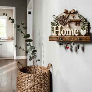 Op de afbeelding: Een houten wand-sleutelhouder met het woord Home in witte letters, een decoratieve bruine sleutel, een jute strik en groene bladaccenten. De plank heeft vier metalen haken met hangende sleutels, geplaatst boven een gevlochten mand.