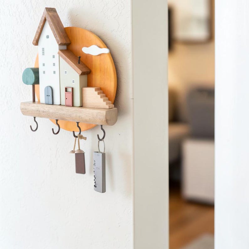 Puede incluir: Un llavero de pared de madera con una escena de casas en miniatura que incluye un edificio blanco, uno verde claro, un &aacute;rbol peque&ntilde;o y una escalera sobre un fondo de sol circular naranja. Cuenta con cuatro ganchos de metal negro para llaves.