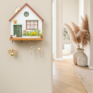 Puede incluir: Un llavero de pared de madera con forma de casa, fachada blanca, techo marr&oacute;n y puerta verde. Incluye un estante con flores amarillas artificiales, un buz&oacute;n y dos figuras de gato. Debajo tiene cuatro ganchos de metal para colgar las llaves.