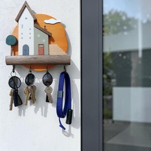 Puede incluir: Un llavero de pared de madera con una escena decorativa de dos casas en blanco y verde salvia sobre un fondo circular naranja. El objeto incluye un estante marr&oacute;n oscuro con cuatro ganchos met&aacute;licos que sostienen varias llaves y un cord&oacute;n azul.