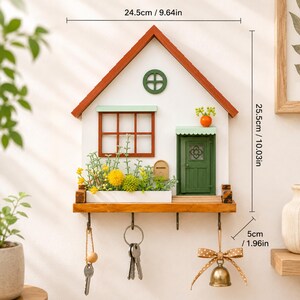 Puede incluir: Un llavero de pared con forma de casa, fachada blanca, techo naranja y puerta verde. Incluye una ventana, una jardinera con flores artificiales amarillas y verdes, y cuatro ganchos met&aacute;licos. Sus dimensiones son 24,5 cm por 25,5 cm por 5 cm.