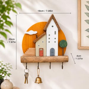 Puede incluir: Un soporte de pared de madera para llaves con dise&ntilde;o de casas sobre un fondo circular amarillo. Incluye cuatro ganchos met&aacute;licos, con llaves y una campana colgadas de dos de ellos. Las dimensiones son 18 cm de ancho, 20 cm de alto y 4 cm de profundidad.