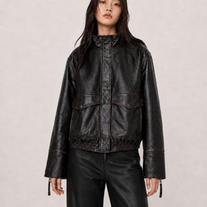 以下が含まれることがあります： ハイカラー、長袖、プラケットで隠されたフロントジッパーを備えた黒のレザージャケット。ジャケットの前面には2つの大きなフラップポケットがあり、ウエストにはギャザー入りの伸縮性のある裾、そして目に見える茶色のステッチのディテールが施されています。