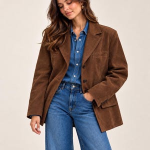 May include: A brown suede blazer featuring a notched lapel, three front buttons, and two flap pockets. The blazer is worn over a blue denim button-down shirt and blue denim jeans. The garment has long sleeves with buttoned cuffs and a structured fit.