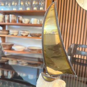 May include: A decorative sailboat sculpture with a gold-coloured sail and a small flag. The sail is framed with a dark metal border and is mounted on a white stone base. The sculpture is displayed on a glass surface.
