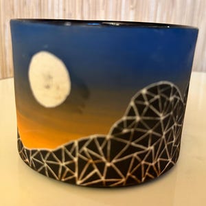 May include: A cylindrical ceramic container with a painted night sky scene. The design features a white moon, an orange and blue gradient sky, and a black geometric mountain range. A wooden ruler is in the background.