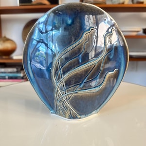 May include: A ceramic vase with a unique, organic shape. The vase features a glossy, blue glaze with darker blue and brown accents. Delicate, linear patterns are etched into the surface, adding texture and visual interest. The vase is displayed on a white surface.