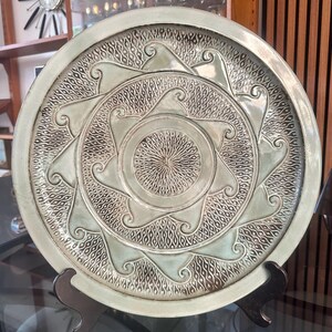 May include: A decorative ceramic plate with a raised, intricate design. The plate features a central sunburst pattern surrounded by stylized waves and a textured background. The color is a muted green-gray, and the plate is displayed on a black stand.