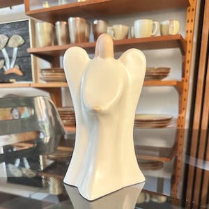 May include: A cream-colored ceramic angel figurine with outstretched wings. The angel has a smooth, glossy finish and a small, rounded head. The figurine is standing on a reflective surface, with shelves of dishware in the background.