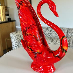 May include: A vibrant red ceramic swan figurine with a glossy finish. The swan has a long, curved neck and outstretched wings, with black and yellow accents. The figurine is a decorative item.