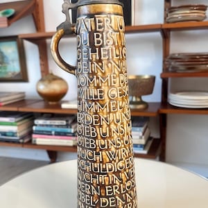 Pode incluir: Um caneco de cerveja de cerâmica vintage com tampa articulada e alça. O caneco é marrom com letras creme. O texto no caneco está em alemão. O caneco tem aproximadamente 30 cm de altura.