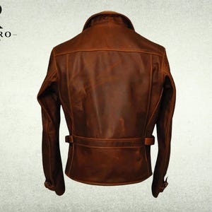 May include: A brown leather jacket shown from the back, featuring a fold-down collar, long sleeves with adjustable cuffs, and a horizontal waist strap with side buckles. The jacket displays a smooth, matte finish with visible seam detailing.