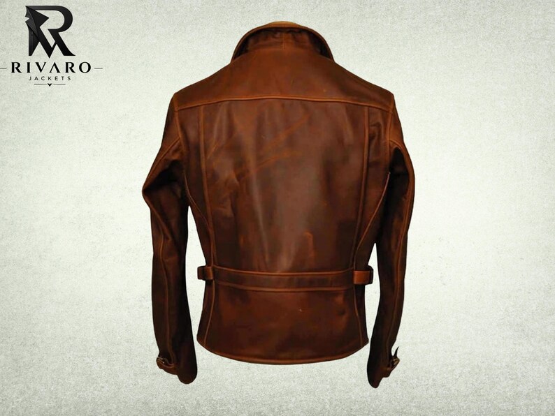 May include: A brown leather jacket shown from the back, featuring a fold-down collar, long sleeves with adjustable cuffs, and a horizontal waist strap with side buckles. The jacket displays a smooth, matte finish with visible seam detailing.