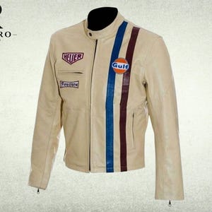 Op de afbeelding: Een beige leren jas met een opstaande kraag, een ritssluiting aan de voorzijde en lange mouwen. De voorzijde is voorzien van een blauwe en donkerrode verticale streep, een rond Gulf-logo en rechthoekige patches met de tekst Heuer en Firestone.