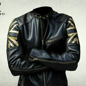 Könnte beinhalten: Eine schwarze Lederjacke mit Stehkragen und Druckknöpfen, einem zentralen Reißverschluss sowie goldfarbenen Union-Jack-Flaggenmustern auf beiden Schultern. Die Ärmel haben Reißverschlussbündchen und die Brust eine diagonale Reißverschlusstasche.