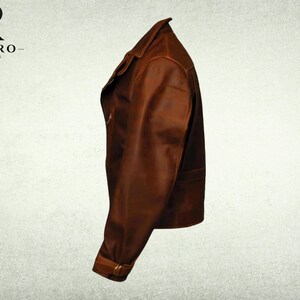 May include: A brown leather jacket shown from a side profile view, featuring a folded collar, a long sleeve with a buckled strap at the cuff, and a smooth finish. The logo for Rivaro Jackets is visible in the upper left corner of the image.
