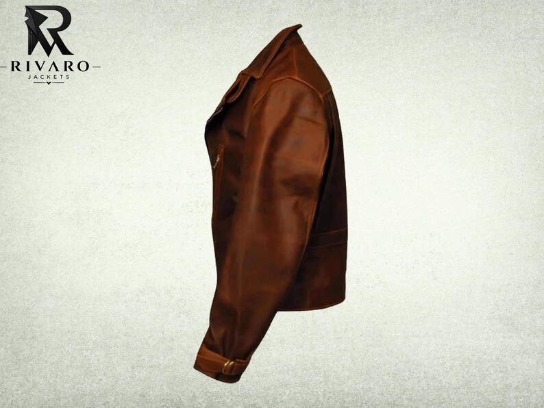 May include: A brown leather jacket shown from a side profile view, featuring a folded collar, a long sleeve with a buckled strap at the cuff, and a smooth finish. The logo for Rivaro Jackets is visible in the upper left corner of the image.