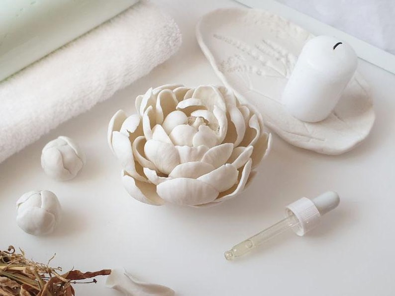 a white flower-shaped object, a white candle, and some dried flowers on a white surface.