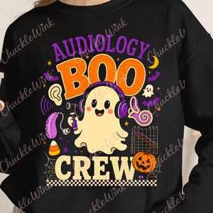 May include: A black long-sleeve sweatshirt featuring a graphic print with the text "AUDIOLOGY BOO CREW". The design includes a ghost wearing purple headphones, a hearing aid, a pumpkin, a candy corn, and an audiogram chart.