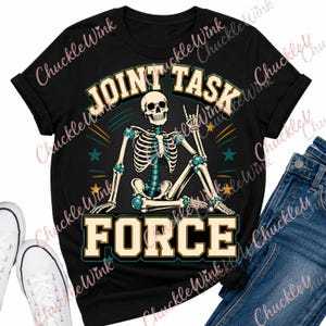 Op de afbeelding: Een zwart t-shirt met een afbeelding van een zittend skelet dat een handgebaar maakt. De tekst "JOINT TASK FORCE" staat in grote, dikgedrukte, lichtgekleurde letters boven en onder het skelet, geaccentueerd door kleine kleurrijke sterren.