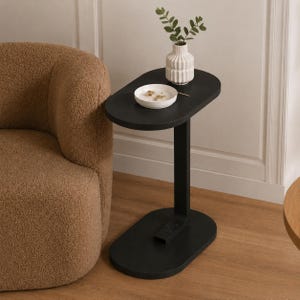 May include: A black side table with an oval top and a single, rectangular support. A small white vase with green sprigs sits on the table, along with a small white dish. The table is next to a brown, textured armchair.