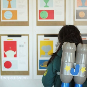Könnte beinhalten: Ein Kind mit einem selbstgemachten Jetpack aus Plastikflaschen, verziert mit Sternen und dem Buchstaben "J". Das Kind steht vor gerahmten Kunstdrucken mit farbenfrohen, minimalistischen Designs.