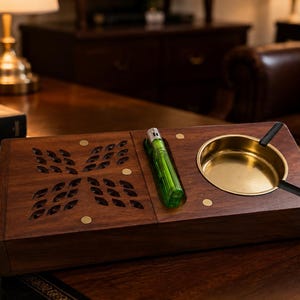 以下が含まれることがあります： 暗い木製の長方形の灰皿。円形の真鍮製ボウル、専用スロットに収められた緑色のプラスチック製ライター、左側に小さな真鍮の円形インレイが施された装飾的な切り抜き模様が特徴です。