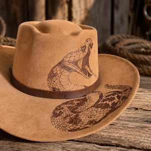 May include: A tan felt cowboy hat featuring a dark brown leather band and a detailed dark brown laser-engraved graphic of a coiled rattlesnake with its mouth open, extending from the crown onto the wide, curved brim of the hat.