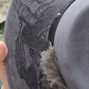 May include: A grey felt hat featuring a detailed black ink illustration of a coiled rattlesnake with fangs bared, accented by a feather and a black leather band with silver-toned metal studs and a decorative circular concho embellishment.