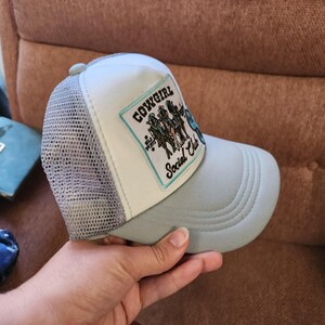 May include: A light grey and white trucker hat featuring a rectangular patch on the front panel. The patch has a light blue border and displays the text "COWGIRL Social Club" in black script and block lettering, accompanied by a graphic of three figures.