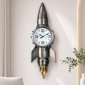 Op de afbeelding: Een wandklok in de vorm van een raket met een metallic grijze afwerking. De wijzerplaat is wit met zwarte cijfers 3, 6, 9 en 12, en zwarte wijzers. De raket heeft puntige vinnen en een goudkleurig vlamontwerp aan de onderkant.