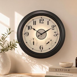 May include: A round wall clock with a black frame and a cream-colored face featuring black Arabic numerals. The clock includes two smaller circular gauges at the bottom for temperature and humidity, along with black hour, minute, and second hands.