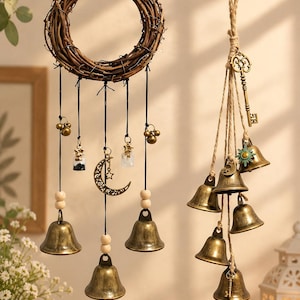 May include: A hanging decoration featuring a circular grapevine wreath with dangling black cords, brass bells, wooden beads, small glass vials, and a crescent moon charm, alongside a separate bundle of brass bells suspended from a vintage key.