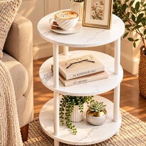 May include: A three-tier round side table with a white faux marble finish and white cylindrical legs. The top shelf holds a coffee cup, a small vase with flowers, and a framed picture. The middle shelf holds two books and glasses.