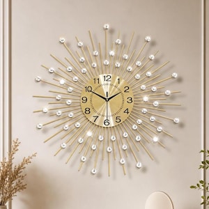 May include: A gold sunburst wall clock featuring a circular face with black Arabic numerals and thin black hands. The clock is surrounded by radiating gold metal spokes, each tipped with a clear, faceted crystal accent that catches the light.