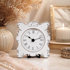 May include: A white table clock with an ornate, scrolled frame and a distressed finish. The circular clock face features black Roman numerals and two black hands. The design includes intricate decorative detailing along the edges of the frame.