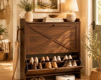 May include: A dark brown wooden shoe storage cabinet with a drop-down door featuring an X-shaped design. The cabinet has an open bottom shelf holding several pairs of shoes, a white robotic vacuum, and a top surface displaying a lamp and books.