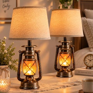 May include: A pair of table lamps designed like vintage oil lanterns with dark bronze-toned metal bases and textured beige fabric drum shades. Each lamp features a central glass chamber containing a warm, glowing flame-effect light source.