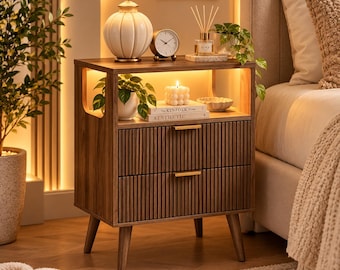 May include: A wooden nightstand with two drawers featuring vertical slats and gold-toned handles. The unit has an open shelf, four tapered legs, and integrated warm lighting. It holds a white lamp, a clock, a candle, and books.