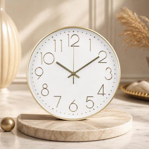 Puede incluir: Un reloj redondo blanco con manecillas doradas y un marco dorado. La esfera del reloj presenta números negros y una pista de minutos punteada. El reloj se muestra sobre un soporte similar al mármol, con una esfera dorada a la izquierda.
