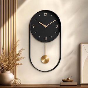 Pode incluir: Um relógio de parede com um mostrador circular preto e numerais dourados nas posições 12, 3, 6 e 9. O relógio possui ponteiros dourados e um pêndulo dourado suspenso dentro de uma estrutura de metal preto de formato oval alongado.