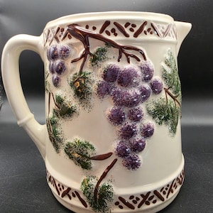 Puede incluir: Una jarra de cerámica de color crema con un diseño en relieve tridimensional de racimos de uvas moradas y hojas verdes sobre vides marrones. Los bordes superior e inferior están decorados con un patrón geométrico de color marrón.
