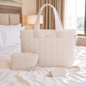 Può includere: Una grande borsa tote trapuntata color bianco sporco con due manici superiori e una pochette rettangolare abbinata con cerniera e cinturino da polso. La borsa presenta un piccolo logo dorato sul davanti e una tracolla regolabile.