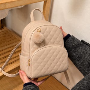 May include: A beige mini backpack featuring a quilted diamond pattern across the front and a small, round, fluffy pom-pom charm attached to the top zip. The bag includes a top handle, adjustable shoulder straps, and a front zipped pocket.