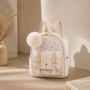 May include: A cream-coloured mini backpack featuring a repeating circular pattern, two beige vertical straps with gold-tone hardware, a top handle, adjustable shoulder straps, and a large, fluffy, cream-coloured pom-pom charm attached to the side.