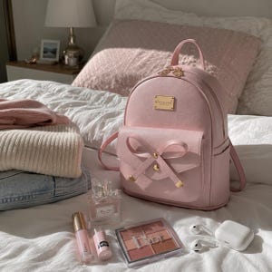 Puede incluir: Una mochila rosa con un lazo decorativo y detalles dorados, colocada sobre una cama. También hay maquillaje, perfume y auriculares inalámbricos, junto con suéteres doblados.