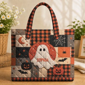May include: A quilted tote bag featuring a patchwork design of black and orange buffalo plaid, grey, and orange squares. The bag displays a central white ghost appliqué with the name SOPHIA, alongside jack-o'-lanterns, bats, and a spider web.