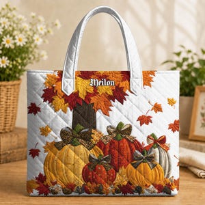 May include: A white quilted tote bag featuring a printed design of four pumpkins in yellow, orange, and red, accented with leopard print bows. The bag displays scattered autumn leaves and the name Meilon in black text. It has two white handles.